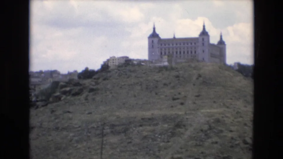 The 1972 Spain footage frames the hilltop castle-like building more centrally, with low buildings or walls nearby and the hillside rising across the foreground.