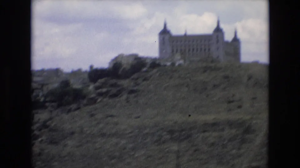 A soft 8mm frame from Spain in 1972 shows the distant castle-like landmark above uneven hillside vegetation and a cloudy sky.