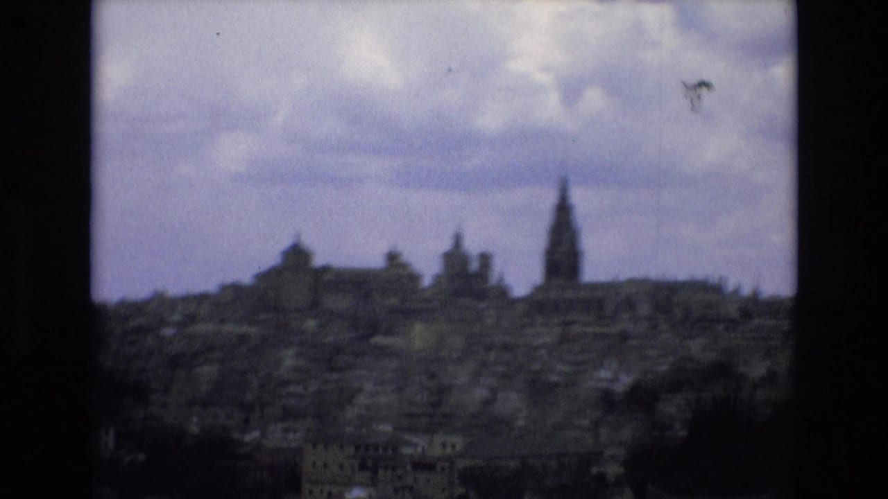 1972 Spain 8mm frame showing a hazy historic cityscape from an elevated vantage point, with clustered buildings and a tall church spire visible against overcast sky.