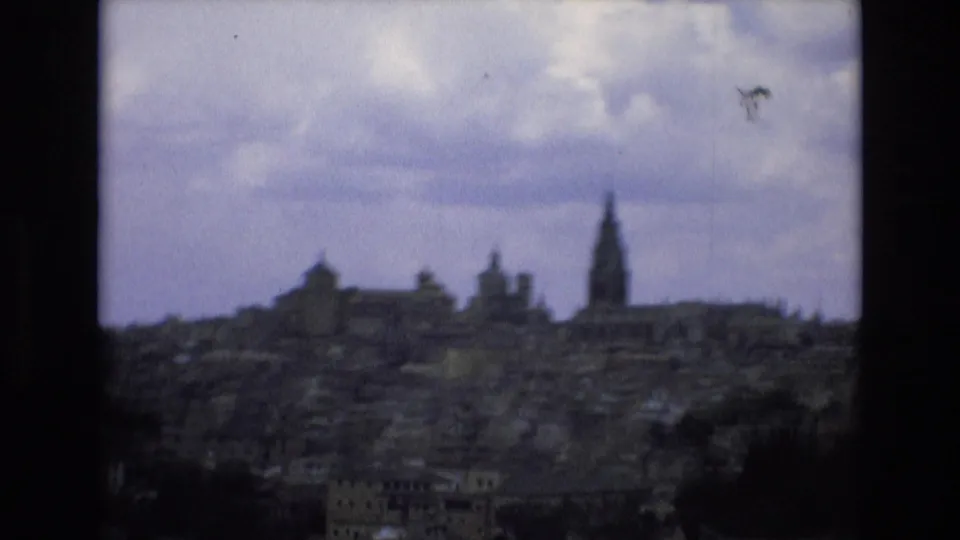 1972 Spain 8mm frame showing a hazy historic cityscape from an elevated vantage point, with clustered buildings and a tall church spire visible against overcast sky.