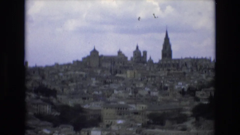 1972 Spain 8mm frame with a clearer wide view of a historic urban hillscape, showing a prominent cathedral-like spire, smaller towers, and dense older buildings.