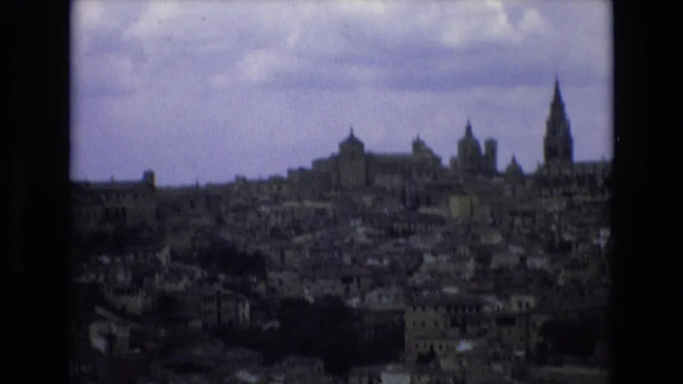 1972 Spain 8mm frame showing a darker distant view of the historic city skyline, with a tall spire at right, church domes or towers, and dense rooftops below a cloudy sky.