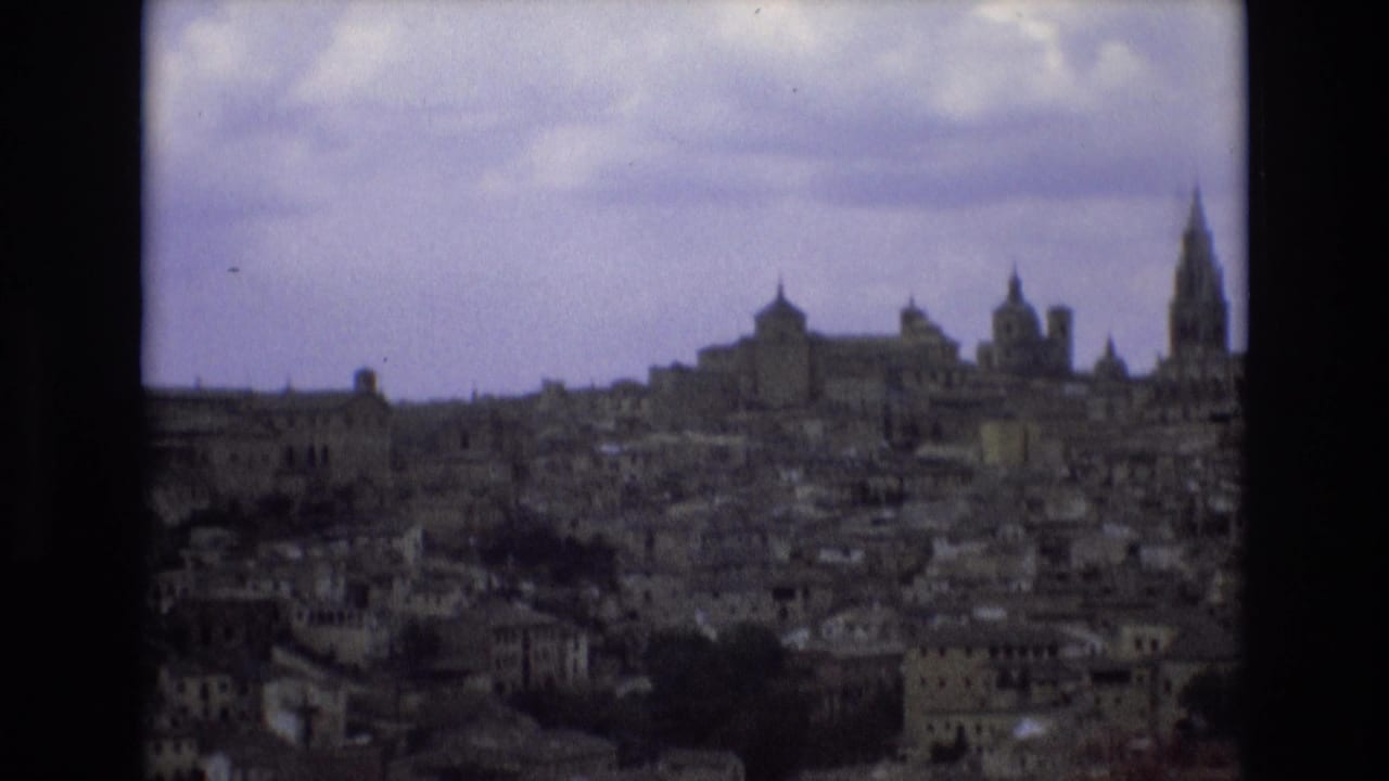 1972 Spain 8mm frame showing a distant historic city skyline with dense buildings, church towers, and a prominent spire under an overcast sky.