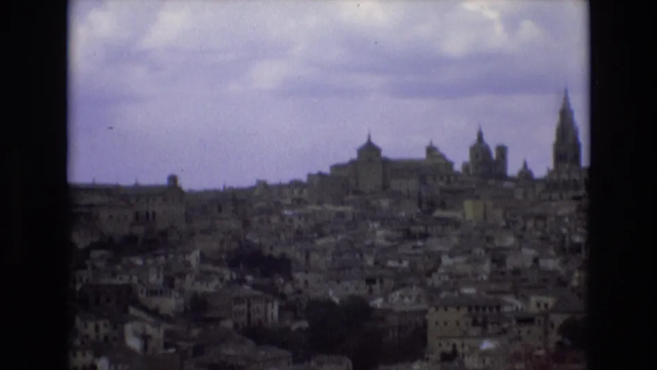 1972 Spain 8mm frame showing a distant historic city skyline with dense buildings, church towers, and a prominent spire under an overcast sky.
