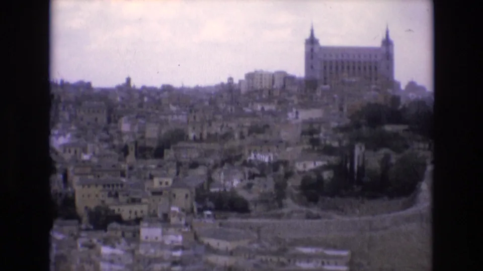 1972 Spain home-movie frame showing a wide hillside cityscape with compact buildings, terraced ground, and a large towered landmark on the ridge.