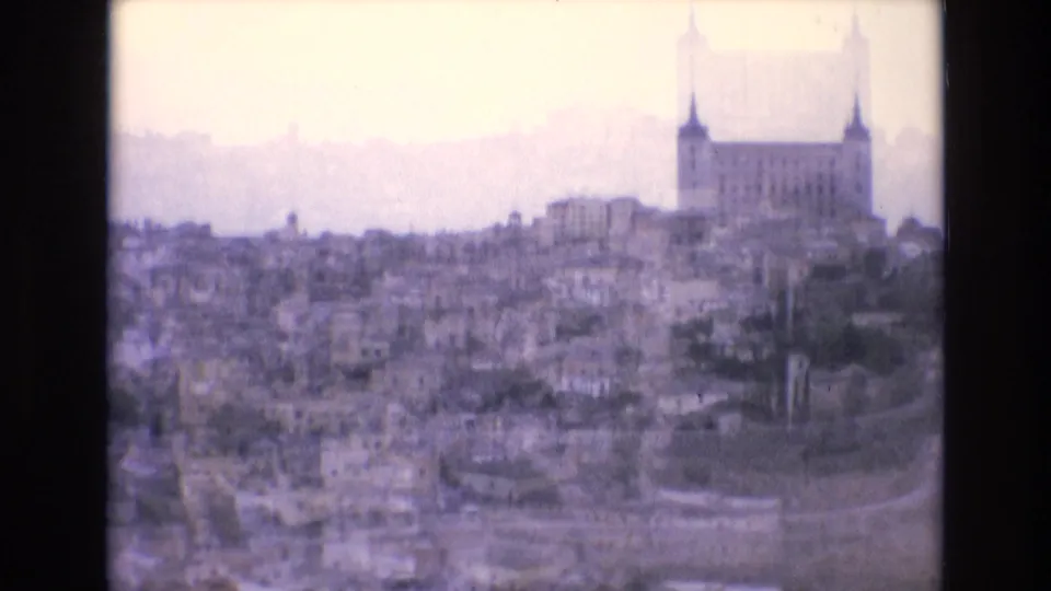 Faded 1972 Spain 8mm frame showing a broad city panorama with dense rooftops and a large towered architectural landmark on the hillside.