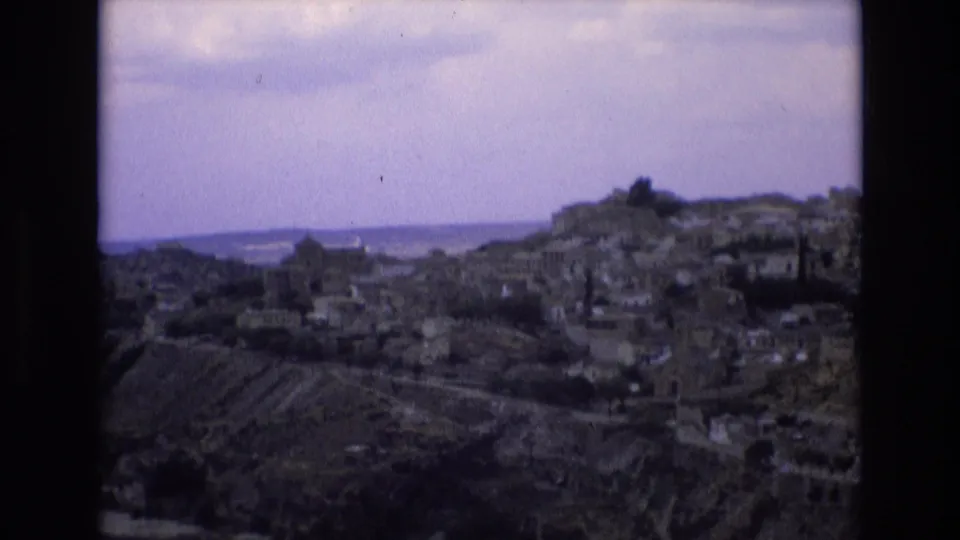 1972 Spain travel footage frame panning across Toledo's hillside neighborhoods, with clustered buildings, terraced slopes, and a pale distant horizon.