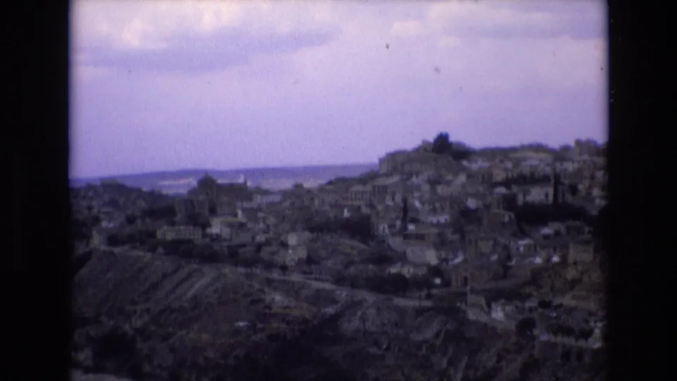 1972 8mm frame from Toledo, Spain, showing hillside buildings, winding roads, terraced ground, and a soft overcast sky during a panoramic tourist view.