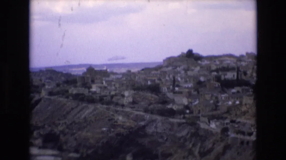 A soft, grainy 8mm view across a Spanish hillside town in 1972, with stone buildings spread over uneven ground beneath a cloudy sky.