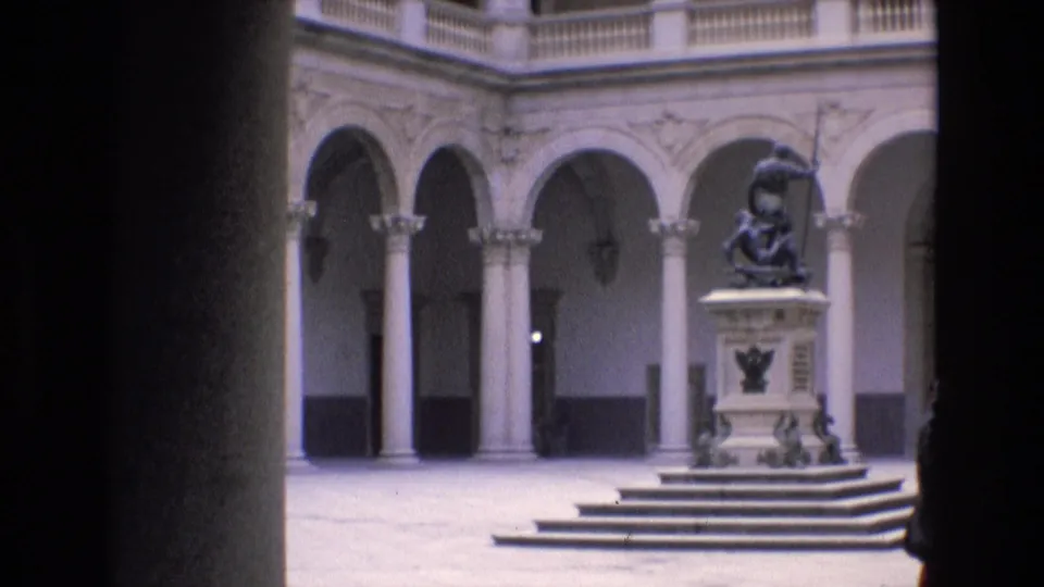 Formal courtyard in Spain with pale arched colonnades, stone columns, upper balustrade, and a central statue raised on a pedestal and steps.