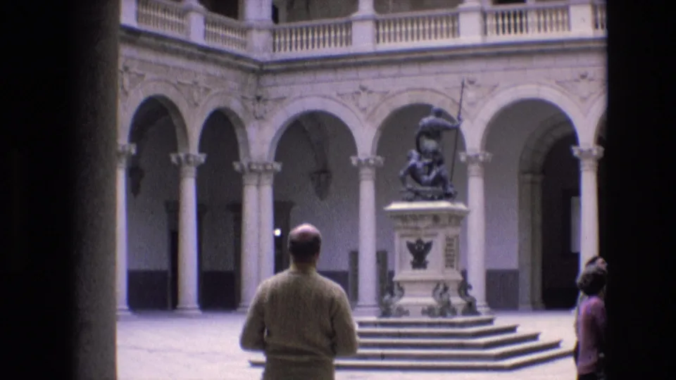 1972 home-movie frame from Spain showing adult visitors in a classical courtyard, with one man seen from behind facing a statue on a pedestal.