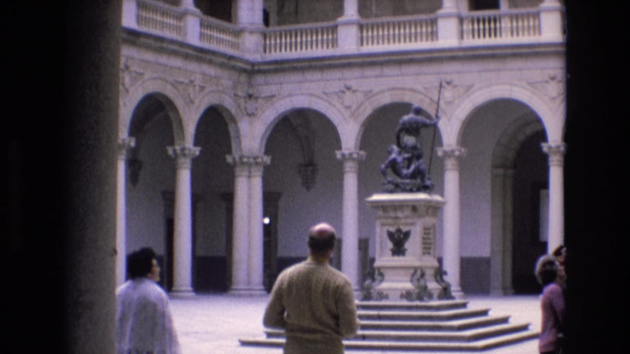 1972 Spain home-movie frame showing visitors in a classical courtyard with arched colonnades, a central stepped pedestal, and a dark spear-holding statue.