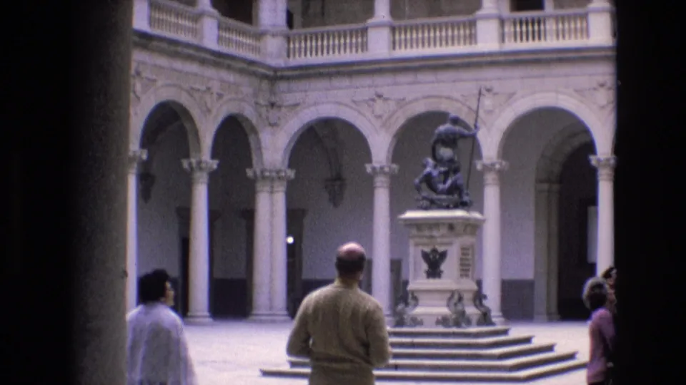 1972 Spain home-movie frame showing visitors in a classical courtyard with arched colonnades, a central stepped pedestal, and a dark spear-holding statue.
