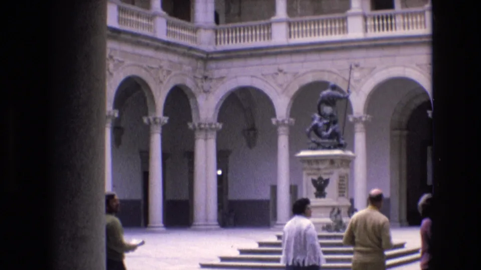 1972 Spain home-movie still of tourists passing through a classical courtyard with a central statue, stone steps, arches, and dark foreground framing.