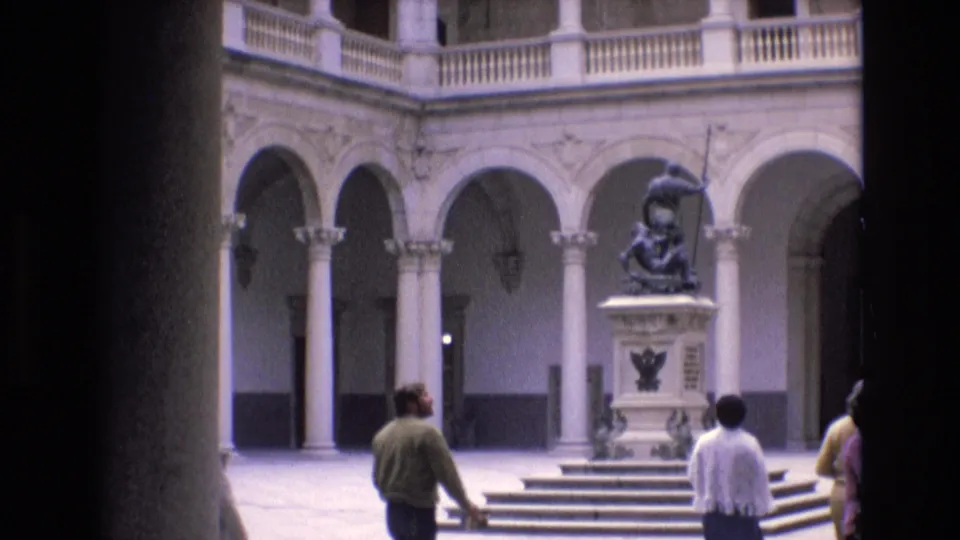 Final 1972 Spain courtyard frame showing visitors walking past the stepped base of a dark spear-bearing statue beneath pale stone arches and columns.