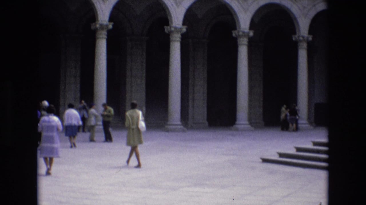 1972 Spain home-movie frame showing tourists in period clothing crossing a paved historic courtyard with tall stone arches and columns in the background.