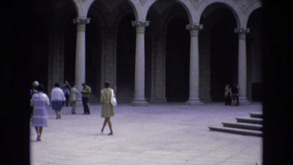 1972 Spain home-movie frame showing tourists in period clothing crossing a paved historic courtyard with tall stone arches and columns in the background.