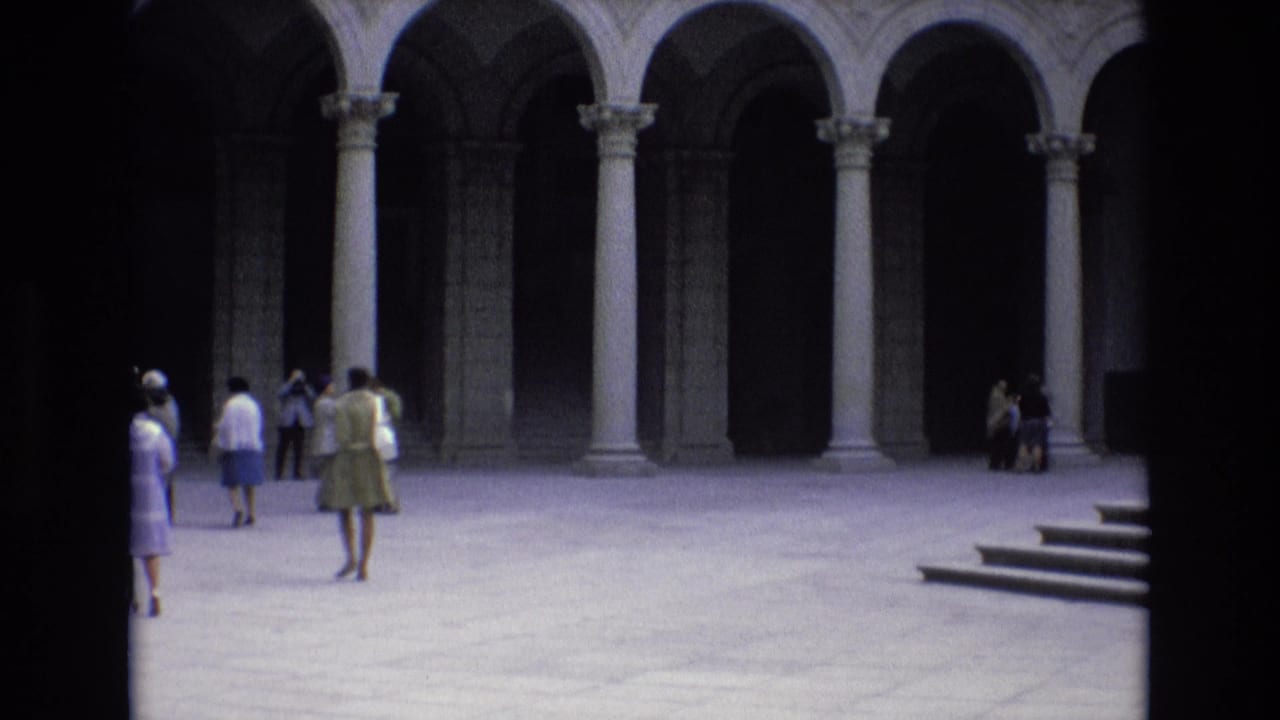 Visitors stand and walk in a sunlit courtyard with stone arches behind them.