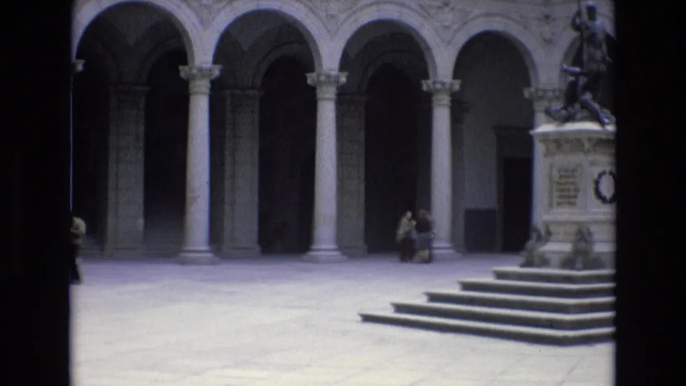 1972 Spain home-movie frame showing a statue on a pedestal with stepped base near arcaded stone architecture and a seated visitor in the background.