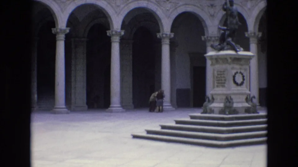 1972 Spain frame of a historic courtyard monument, with a raised statue base, broad steps, arched stone openings, and a seated visitor near the arcade.