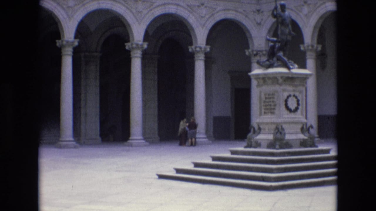 1972 Spain 8mm frame showing a historic stone courtyard with arched colonnades, a raised monument topped by a statue, and small visitor figures near the base.