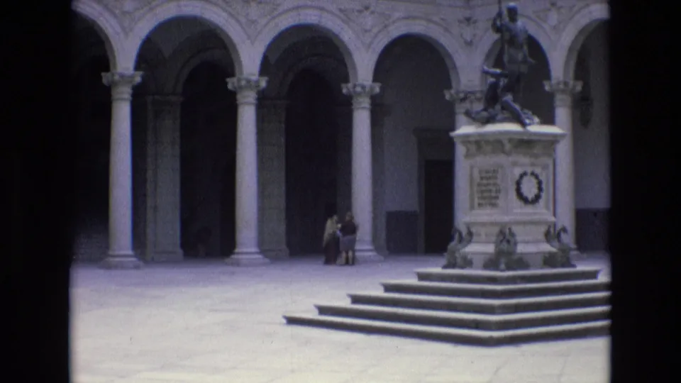 1972 Spain 8mm frame showing a historic stone courtyard with arched colonnades, a raised monument topped by a statue, and small visitor figures near the base.
