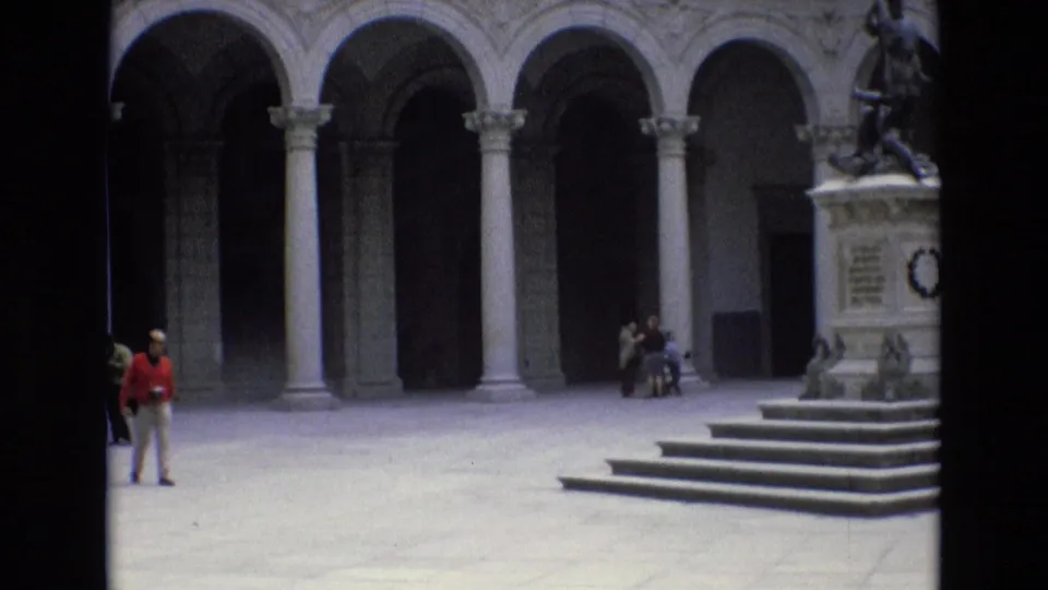 1972 Spain 8mm frame showing a person in a red shirt crossing a sunlit courtyard, with the monument, steps, and arched stone colonnade behind them.