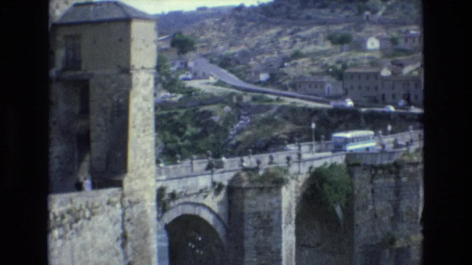 1972 Spain travel frame showing a stone bridge spanning a valley, a white bus on the road, hillside buildings, and a winding route through rugged terrain.