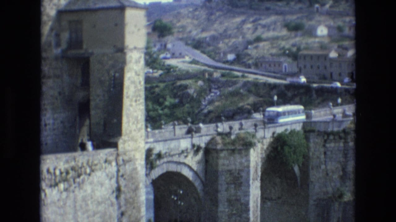 1972 Spain 8mm frame showing a historic stone bridge with arched supports, hillside buildings, pedestrians, and a white bus crossing the roadway.