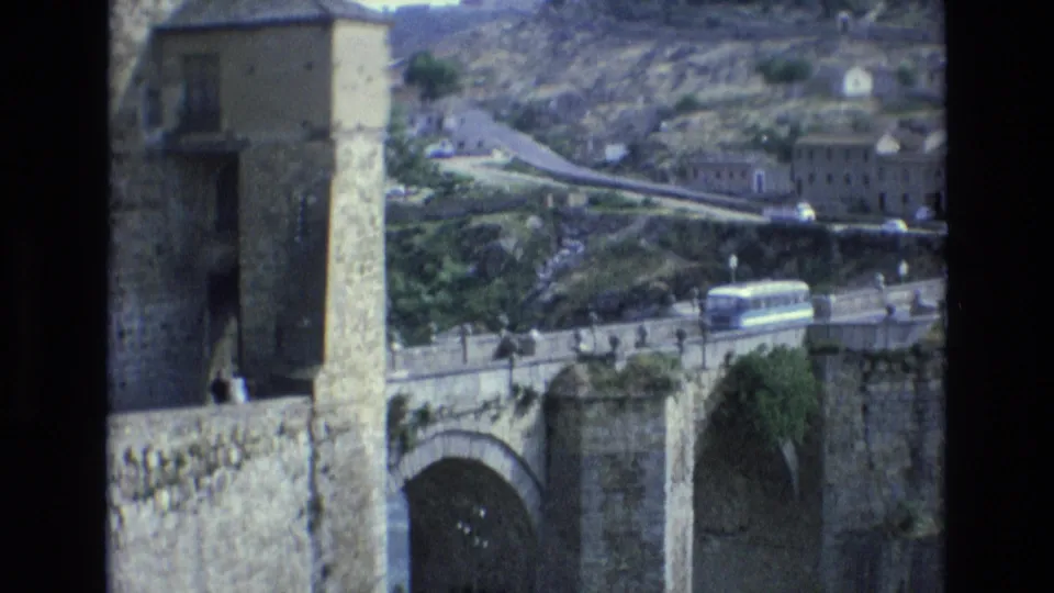 1972 Spain 8mm frame showing a historic stone bridge with arched supports, hillside buildings, pedestrians, and a white bus crossing the roadway.