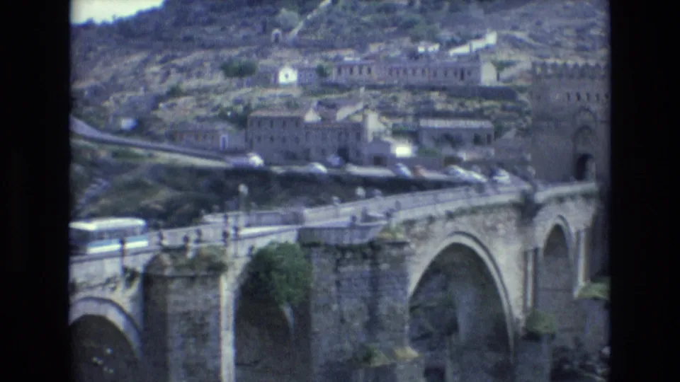 1972 Spain 8mm frame with a multi-arch stone bridge, people along the bridge path, a road, and masonry buildings on the hillside.