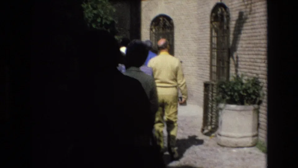 1972 Spain 8mm frame showing pedestrians in a shaded cobblestone alley, including a man in a yellow outfit near brick walls and barred arched windows.