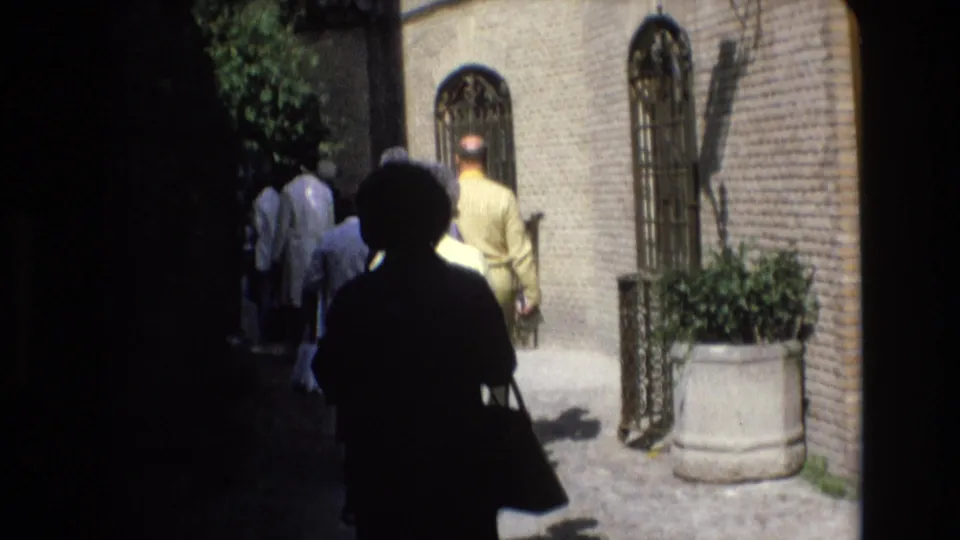 1972 Spain 8mm frame with silhouetted figures entering a sunlit cobblestone alley beside brick walls, arched windows, and a large planter.
