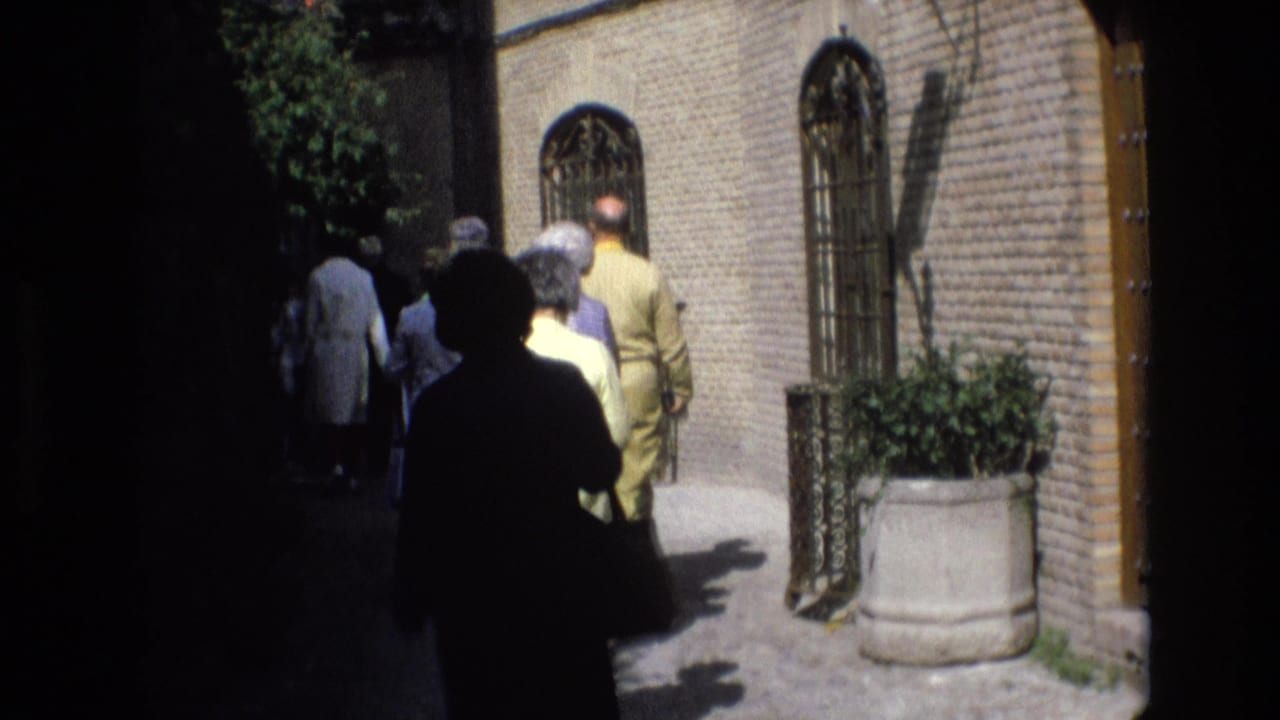 1972 Spain home-movie frame showing pedestrians walking through a shaded historic street with brick walls, iron gates, and a large stone planter.
