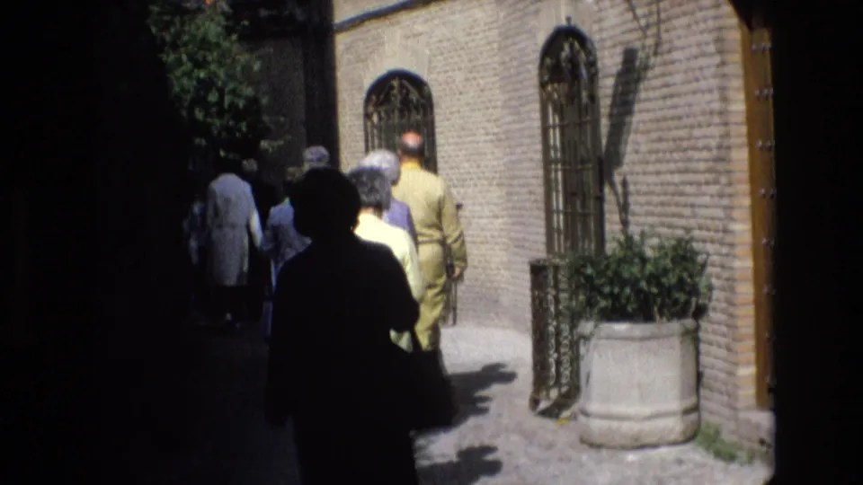 1972 Spain home-movie frame showing pedestrians walking through a shaded historic street with brick walls, iron gates, and a large stone planter.