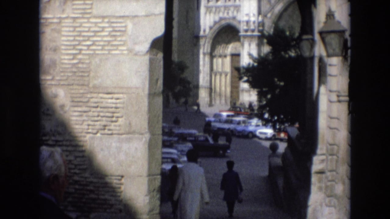 View through an archway toward an ornate facade, pedestrians, and cars on a street.