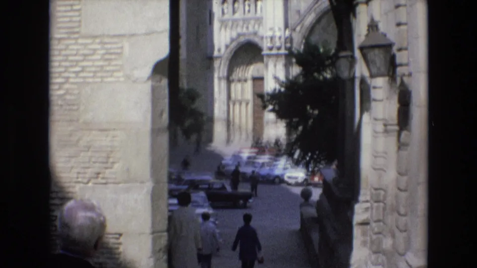 1972 Spain frame showing a street scene beyond stone walls, with pedestrians, 1970s cars, a lamp, and ornate historic architectural detail.