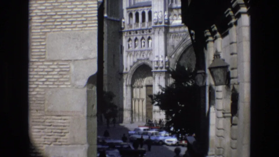 1972 Spain home-movie frame with a shaded passageway view of an ornate historic facade, street lamps, pedestrians, and parked cars.