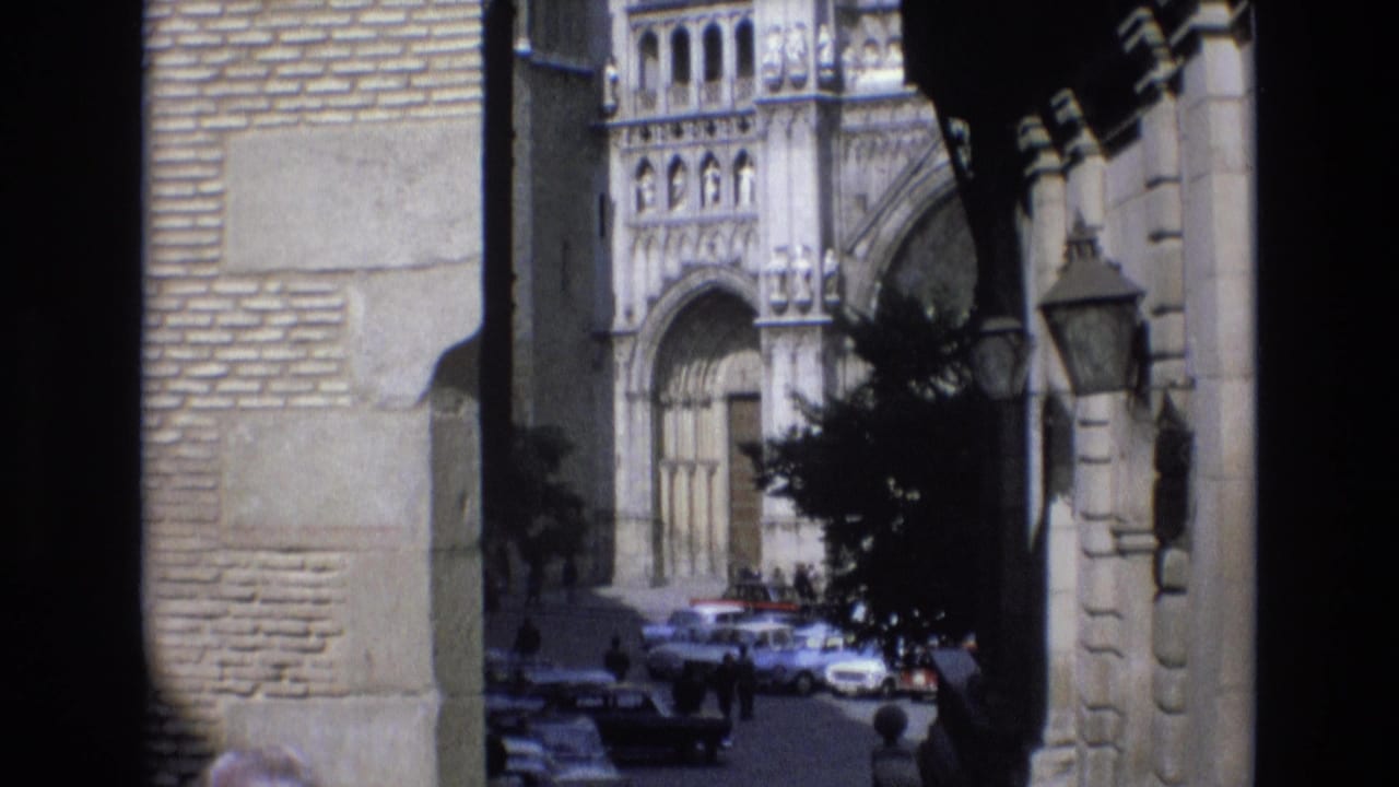 1972 Spain 8mm frame looking from a shaded stone arch toward a Gothic-style cathedral entrance, parked cars, pedestrians, and a hanging street lantern.