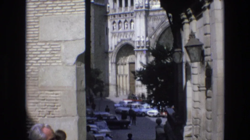1972 Spain street scene framed by dark arch columns, showing a cathedral facade, parked cars, small pedestrian figures, and sunlit stonework.