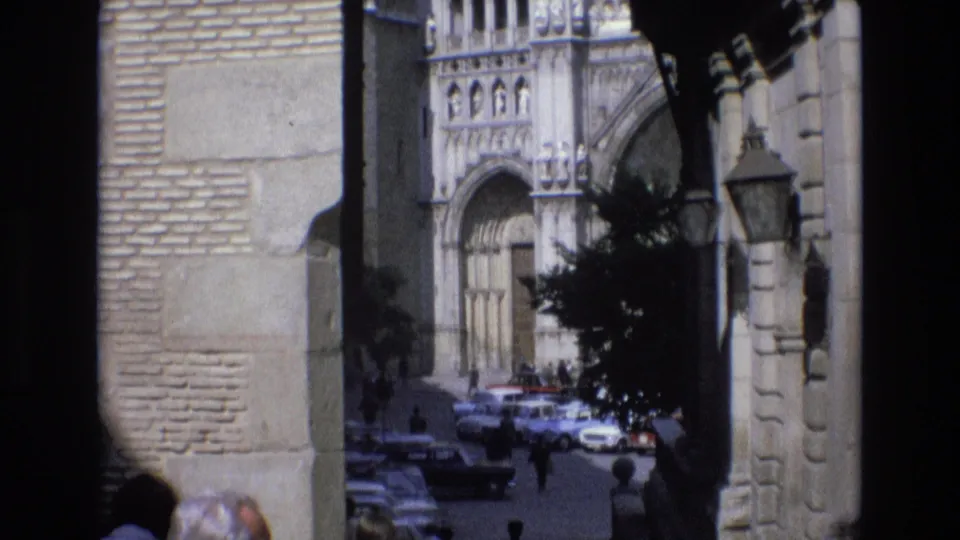 1972 Spain 8mm frame from beneath stone arches, with cathedral stonework in the background, 1970s parked automobiles, pedestrians, and foreground heads at the lower edge.