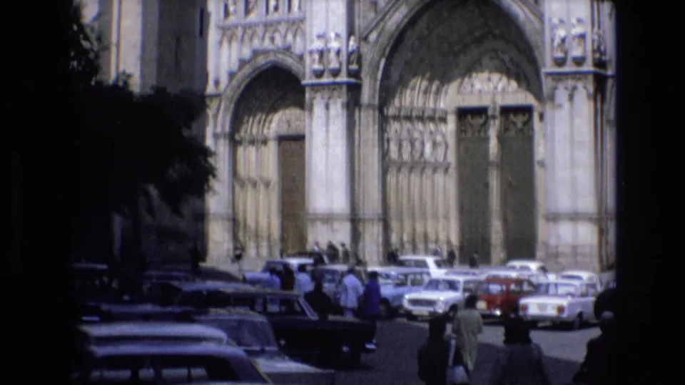 Closer 1972 Spain 8mm view of a Gothic-style cathedral doorway, a row of parked cars, pedestrians crossing the street, and deep shadow from nearby trees or arches.