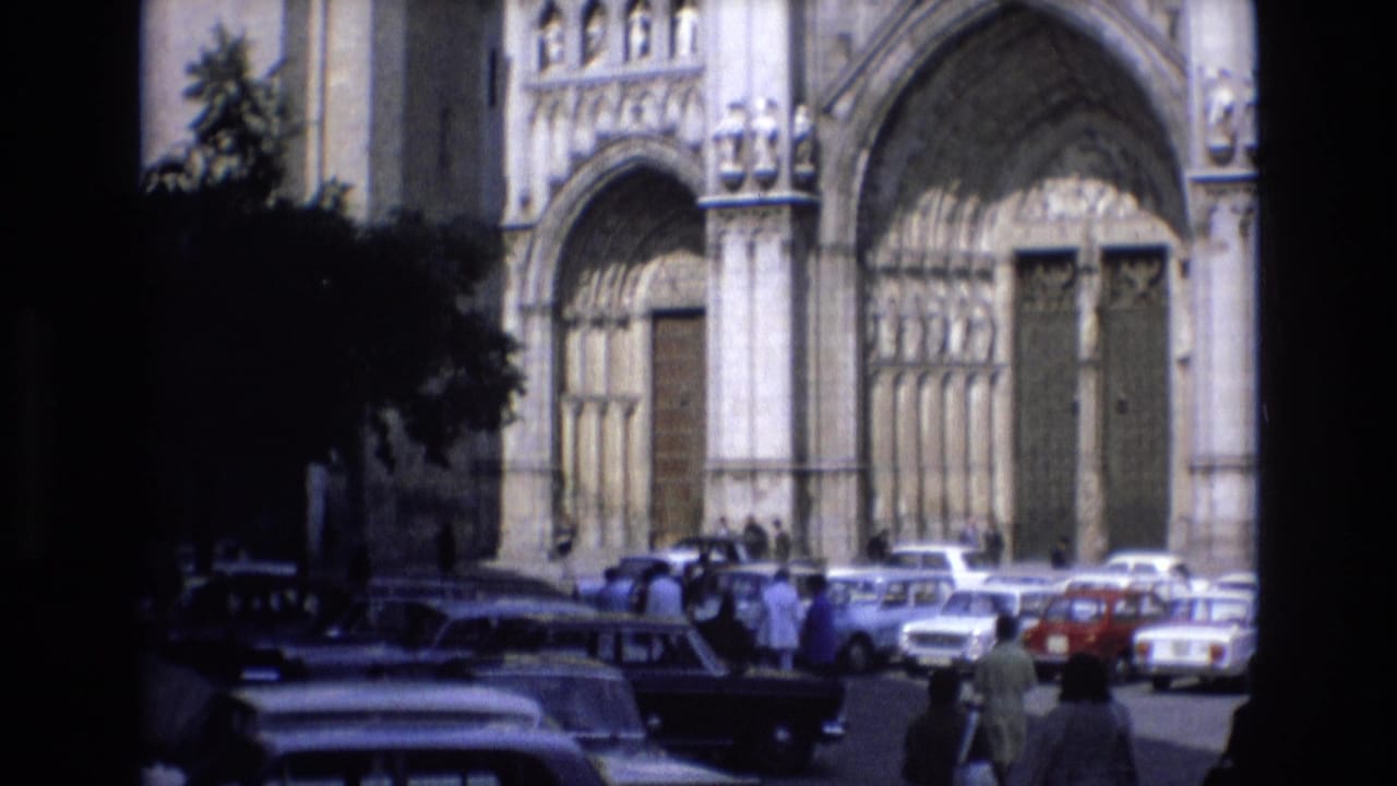 1972 Spain home-movie frame showing people walking near parked sedans in front of a grand stone cathedral facade with arched doors.