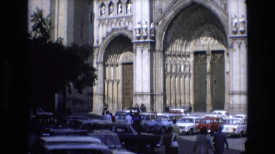 Home-movie view of pedestrians moving past classic sedans toward a large stone cathedral entrance in Spain, dated by metadata to 1972.