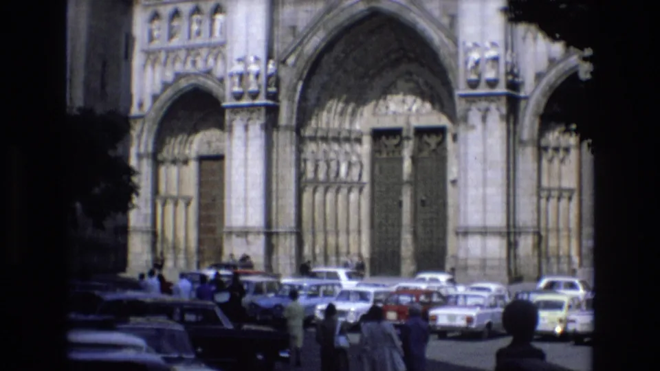 Final 1972 Spain frame with a Gothic-style cathedral entrance, rows of parked cars, and pedestrians partly framed by deep shadow and foliage.