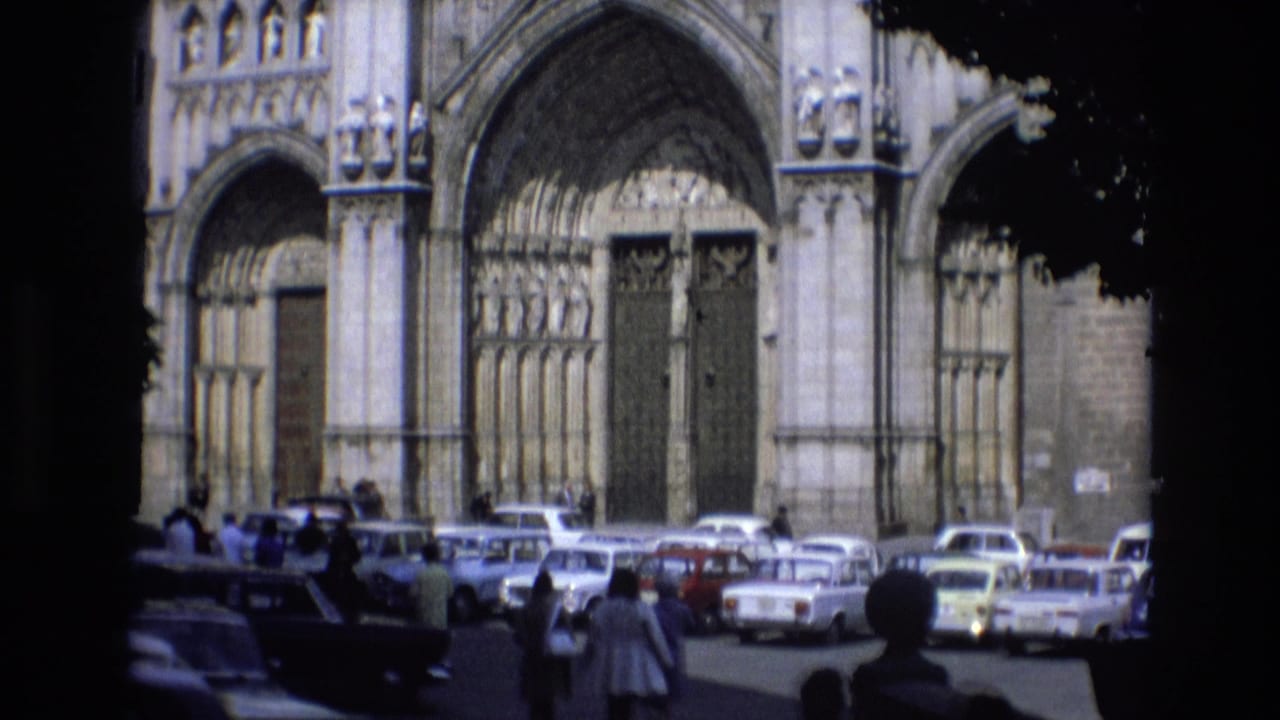 1972 Spain home-movie frame showing a Gothic cathedral facade beyond a busy parking lot, with pedestrians and period cars seen from shade or foliage.