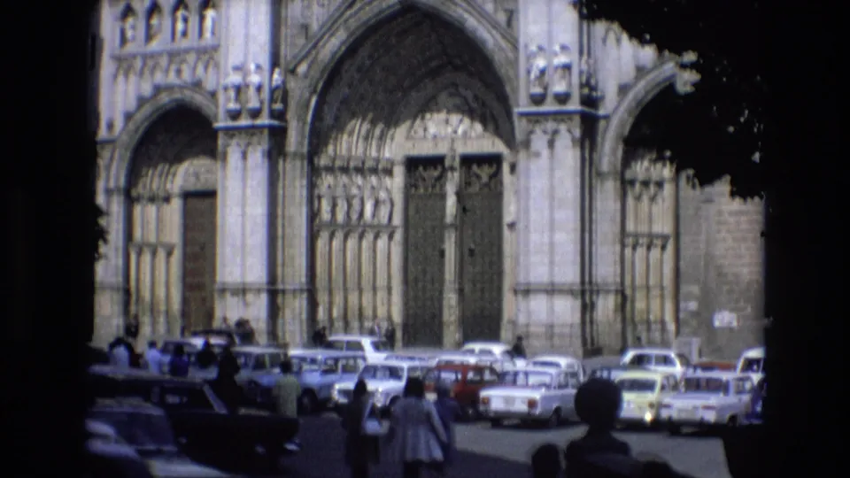 1972 Spain home-movie frame showing a Gothic cathedral facade beyond a busy parking lot, with pedestrians and period cars seen from shade or foliage.