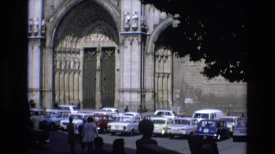 1972 Spain frame of a Gothic cathedral exterior with carved arches, multiple parked European cars, and small figures walking through the foreground lot.