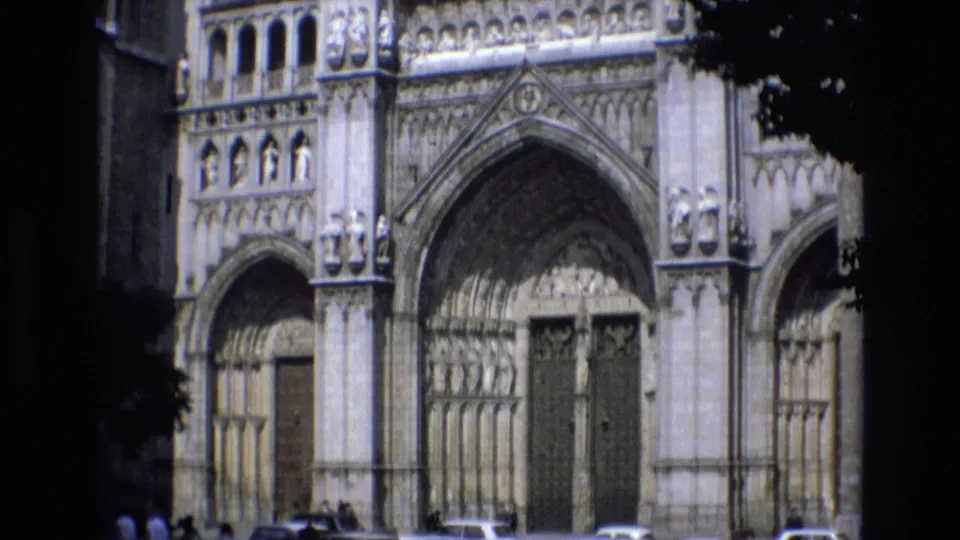 Spain 1972 frame of the cathedral exterior, with ornate stone entrances above a street edge where classic cars and small pedestrians are visible.