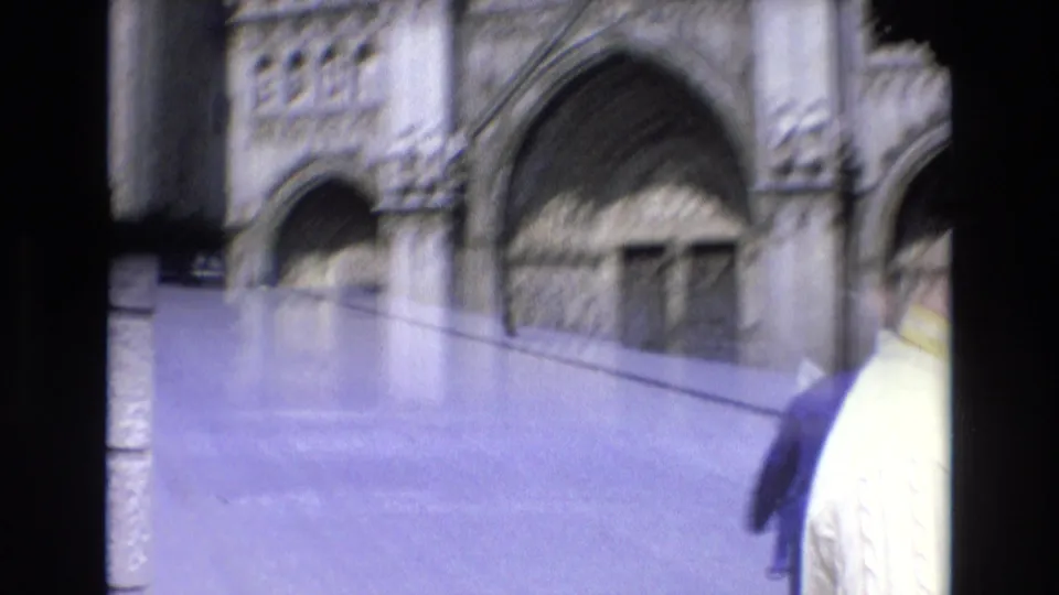 Spain 1972 home-movie frame with a soft, handheld view of the Gothic facade across an open paved area and partially visible pedestrians in the foreground.